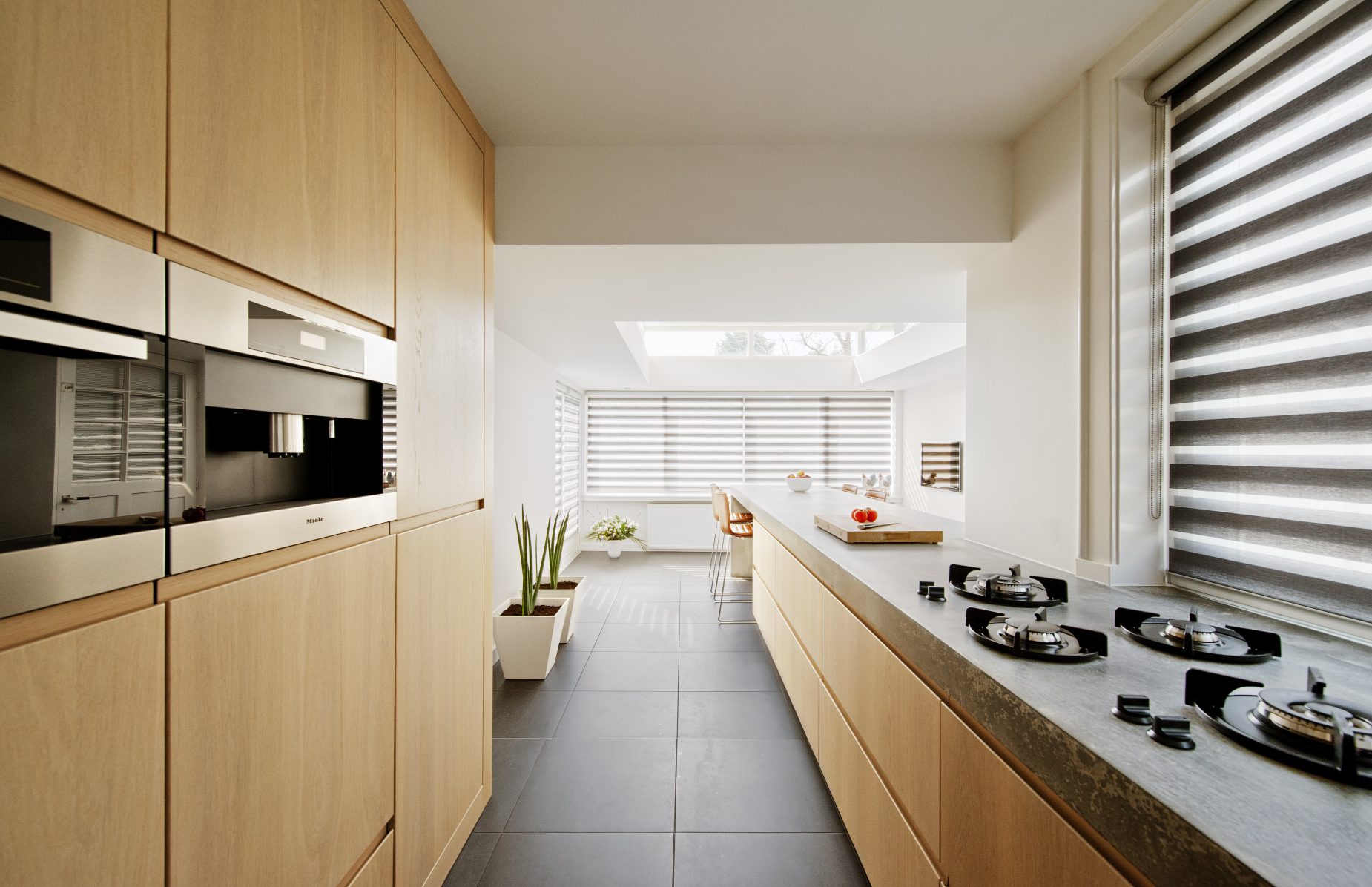 Villa-Naarden-1 Wij hebben een nieuw ontwerp gemaakt voor de keuken en bijkeuken met een uitbouw. Om de huidige keukenruimte te betrekken bij de uitbouw hebben wij het aanrecht als verbindende element gemaakt. Het bijna 8 meter lange aanrechtblad is gemaakt van beton en is op locatie (in het werk) gestort en gepolijst. Het betonnen aanrecht bevat geïntegreerd ‘pitt cooking’. De kasten van de keuken zijn gemaakt van licht eiken. Het aanrechtblad loopt over naar een bargedeelte. Bij het bargedeelte zit in het betonnen aanrecht plaatselijk een elektrische ‘vloerverwarming’, op deze manier is de ‘kou eraf’ als men aan de bar zit. Boven het bargedeelte is het dak plaatselijk verhoogd met daarin ramen rondom. Door deze ramen komt een mooie natuurlijke lichtinval in het eetgedeelte.