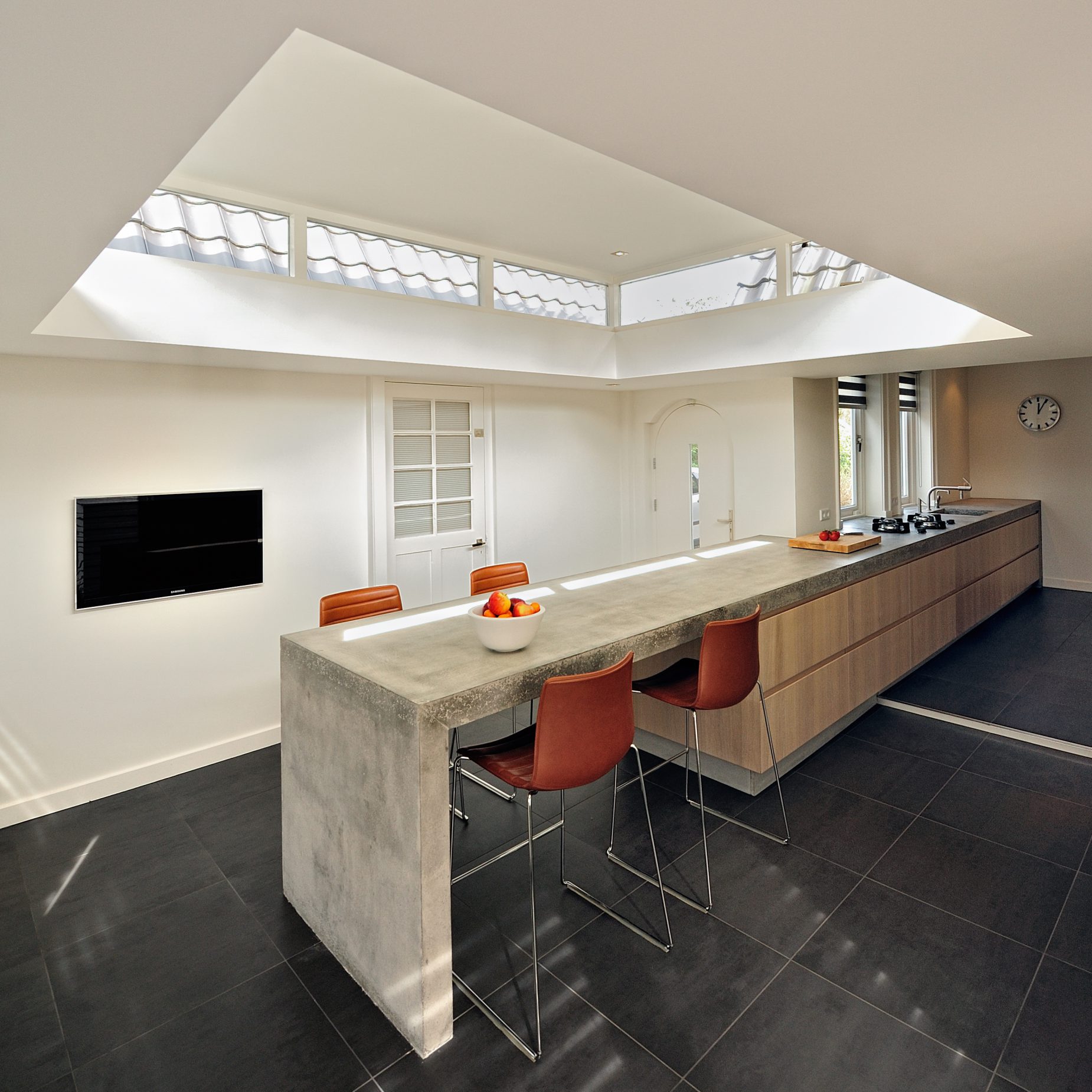 Villa-Naarden-2 Wij hebben een nieuw ontwerp gemaakt voor de keuken en bijkeuken met een uitbouw. Om de huidige keukenruimte te betrekken bij de uitbouw hebben wij het aanrecht als verbindende element gemaakt. Het bijna 8 meter lange aanrechtblad is gemaakt van beton en is op locatie (in het werk) gestort en gepolijst. Het betonnen aanrecht bevat geïntegreerd ‘pitt cooking’. De kasten van de keuken zijn gemaakt van licht eiken. Het aanrechtblad loopt over naar een bargedeelte. Bij het bargedeelte zit in het betonnen aanrecht plaatselijk een elektrische ‘vloerverwarming’, op deze manier is de ‘kou eraf’ als men aan de bar zit. Boven het bargedeelte is het dak plaatselijk verhoogd met daarin ramen rondom. Door deze ramen komt een mooie natuurlijke lichtinval in het eetgedeelte.