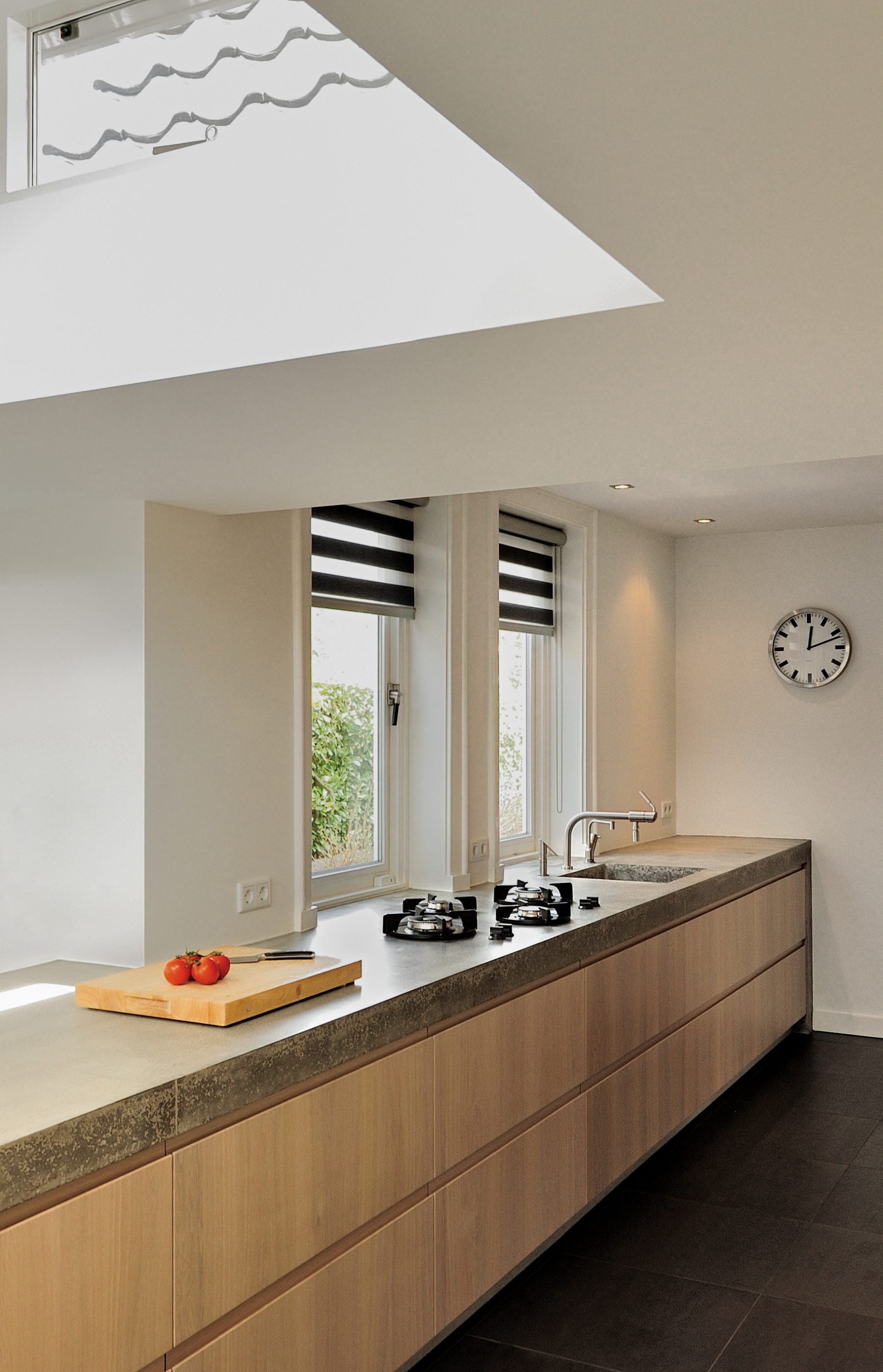 Villa-Naarden-3 Wij hebben een nieuw ontwerp gemaakt voor de keuken en bijkeuken met een uitbouw. Om de huidige keukenruimte te betrekken bij de uitbouw hebben wij het aanrecht als verbindende element gemaakt. Het bijna 8 meter lange aanrechtblad is gemaakt van beton en is op locatie (in het werk) gestort en gepolijst. Het betonnen aanrecht bevat geïntegreerd ‘pitt cooking’. De kasten van de keuken zijn gemaakt van licht eiken. Het aanrechtblad loopt over naar een bargedeelte. Bij het bargedeelte zit in het betonnen aanrecht plaatselijk een elektrische ‘vloerverwarming’, op deze manier is de ‘kou eraf’ als men aan de bar zit. Boven het bargedeelte is het dak plaatselijk verhoogd met daarin ramen rondom. Door deze ramen komt een mooie natuurlijke lichtinval in het eetgedeelte.