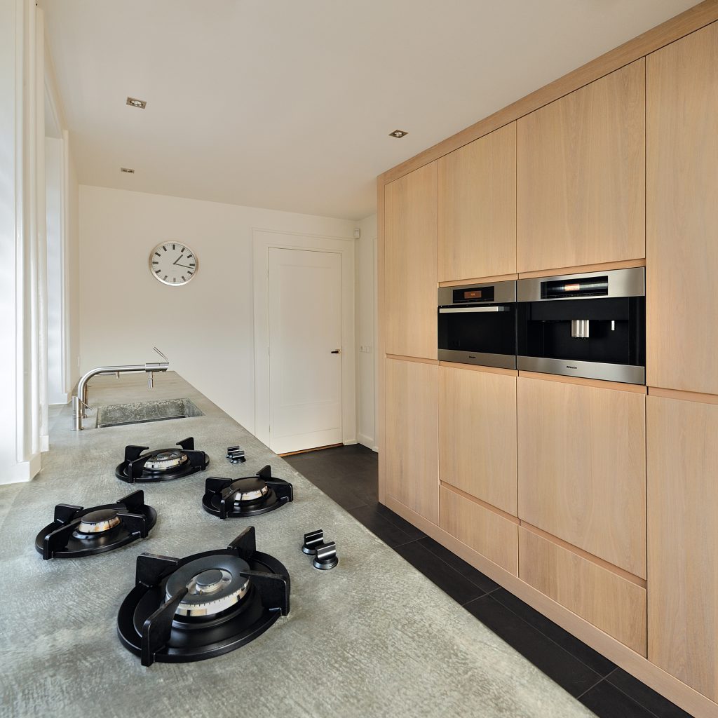 Villa-Naarden-4 Wij hebben een nieuw ontwerp gemaakt voor de keuken en bijkeuken met een uitbouw. Om de huidige keukenruimte te betrekken bij de uitbouw hebben wij het aanrecht als verbindende element gemaakt. Het bijna 8 meter lange aanrechtblad is gemaakt van beton en is op locatie (in het werk) gestort en gepolijst. Het betonnen aanrecht bevat geïntegreerd ‘pitt cooking’. De kasten van de keuken zijn gemaakt van licht eiken. Het aanrechtblad loopt over naar een bargedeelte. Bij het bargedeelte zit in het betonnen aanrecht plaatselijk een elektrische ‘vloerverwarming’, op deze manier is de ‘kou eraf’ als men aan de bar zit. Boven het bargedeelte is het dak plaatselijk verhoogd met daarin ramen rondom. Door deze ramen komt een mooie natuurlijke lichtinval in het eetgedeelte.