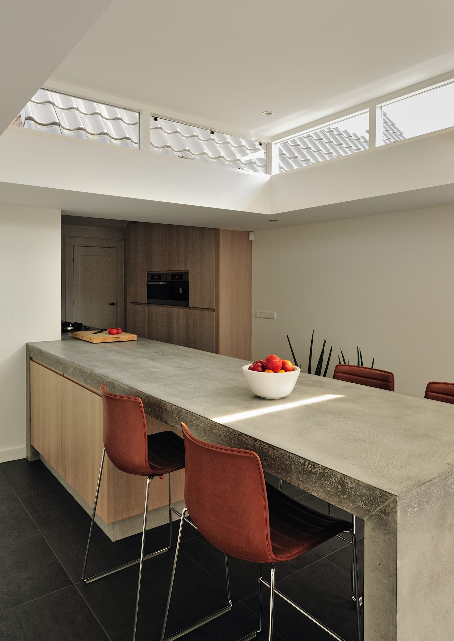 Villa-Naarden-6 Wij hebben een nieuw ontwerp gemaakt voor de keuken en bijkeuken met een uitbouw. Om de huidige keukenruimte te betrekken bij de uitbouw hebben wij het aanrecht als verbindende element gemaakt. Het bijna 8 meter lange aanrechtblad is gemaakt van beton en is op locatie (in het werk) gestort en gepolijst. Het betonnen aanrecht bevat geïntegreerd ‘pitt cooking’. De kasten van de keuken zijn gemaakt van licht eiken. Het aanrechtblad loopt over naar een bargedeelte. Bij het bargedeelte zit in het betonnen aanrecht plaatselijk een elektrische ‘vloerverwarming’, op deze manier is de ‘kou eraf’ als men aan de bar zit. Boven het bargedeelte is het dak plaatselijk verhoogd met daarin ramen rondom. Door deze ramen komt een mooie natuurlijke lichtinval in het eetgedeelte.